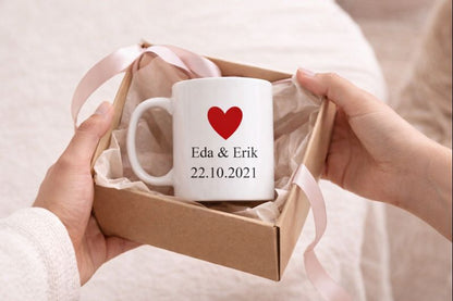Person hält eine Geschenkbox mit einer weißen personalisierten Tasse, verziert mit rotem Herz und den Namen Eda & Erik sowie dem Datum 22.10.2021.